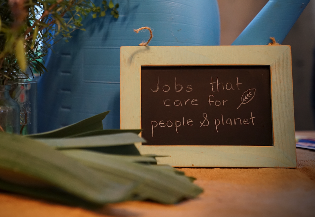 A small chalkboard with light wooden frame reads "Jobs that care for people & planet" with a leaf illustration. It sits on a table next to green leaves, a glass jar of herbs, and a large blue watering can in the background.